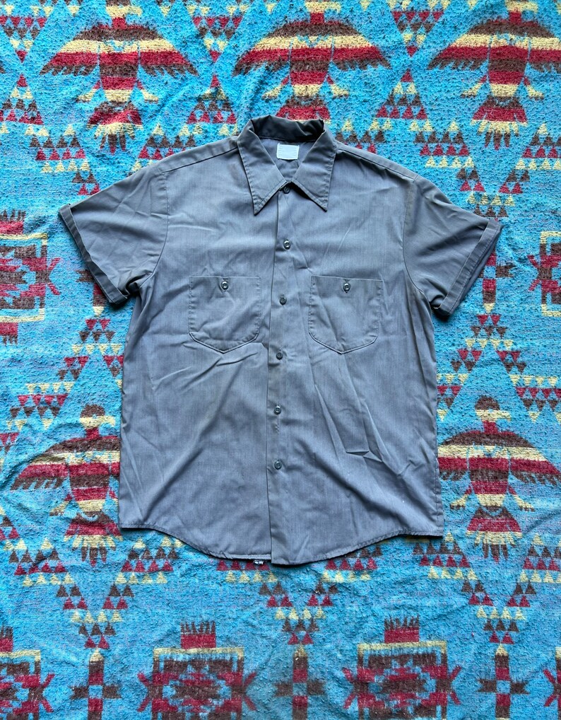 以下が含まれることがあります： ボタンダウンカラーと胸ポケットが2つ付いたグレーの半袖ワークシャツです。