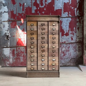 May include: A vintage wooden cabinet with multiple drawers, each with a small label. The cabinet is in a worn condition, with some of the paint chipped away. The cabinet is standing in front of a red and gray wall.
