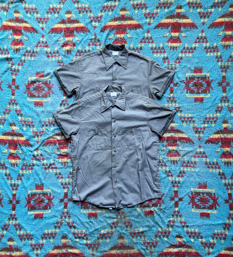 以下が含まれることがあります： 2枚のグレーの半袖シャツが重ねられています。シャツはボタンダウンの前立てと尖った襟が付いています。