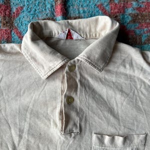 May include: A light beige polo shirt with a small white tag on the collar and a single pocket on the left side. The shirt has a button-up closure.