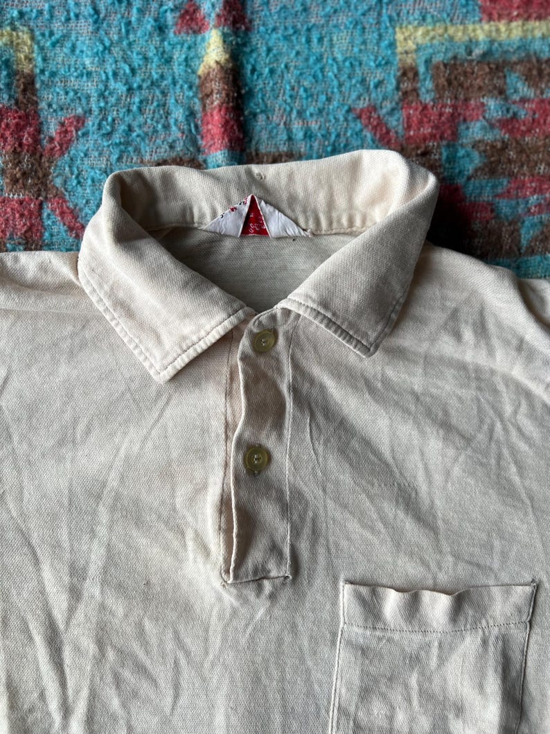 May include: A light beige polo shirt with a small white tag on the collar and a single pocket on the left side. The shirt has a button-up closure.
