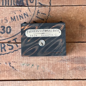 May include: A black wooden box with a metal plaque that reads "Farmers National Bank 1478 Princeton, Ill.". The box has a keyhole and is sitting on a wooden surface.