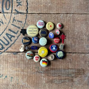 May include: A collection of vintage button pins with various designs and colors, including flags, logos, and slogans. The pins are arranged in a heart shape on a wooden surface.