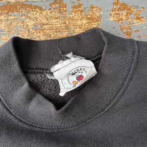Puede incluir: Sudadera negra con una etiqueta de logotipo de Mickey Mouse. La etiqueta es blanca con contornos negros y una cabeza de Mickey Mouse roja.