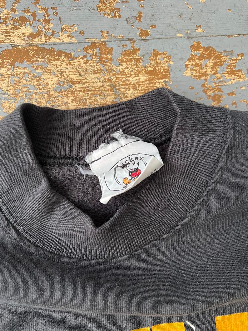 Puede incluir: Sudadera negra con una etiqueta de logotipo de Mickey Mouse. La etiqueta es blanca con contornos negros y una cabeza de Mickey Mouse roja.