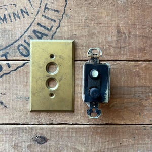 May include: A vintage brass light switch cover with two circular openings, next to a black and white switch mechanism. The items rest on a weathered wooden surface, showcasing an antique aesthetic.
