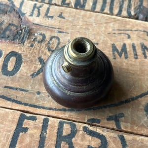 May include: A dark brown, vintage-style knob with a brass-colored threaded top. The knob has a rounded shape and a small screw on the side. The background is a wooden surface with faded black lettering.