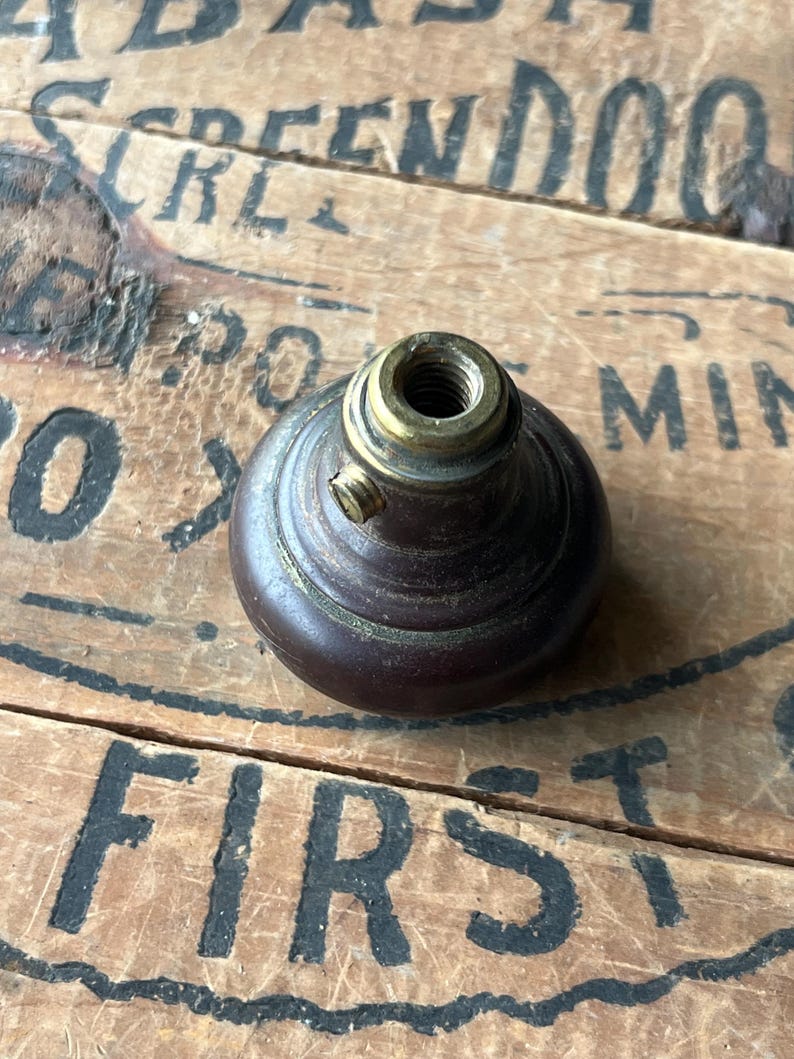 May include: A dark brown, vintage-style knob with a brass-colored threaded top. The knob has a rounded shape and a small screw on the side. The background is a wooden surface with faded black lettering.