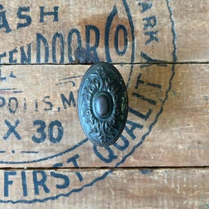 May include: A dark green oval-shaped door knob with intricate floral designs and a circular center piece. The knob is on a wooden surface with black text.