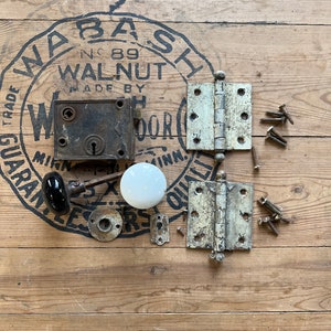 May include: Antique door hardware set including a door knob, a door lock, two hinges, and screws. The hardware is made of metal and has a worn, aged finish. The door knob is black with a white porcelain insert. The door lock is rusty and has a keyhole. The hinges are white with a brass finish. The screws are silver.