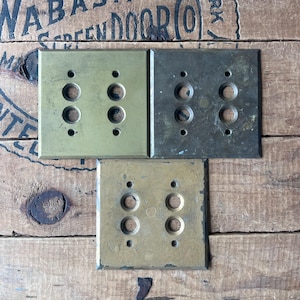 May include: Three vintage brass light switch plates with two sets of holes each. The plates are rectangular and have a slightly aged appearance. The background is a wooden surface with the text "ORIENTAL MADE BY WABASH SCREEN DOOR (O)" visible.