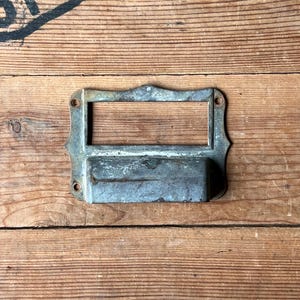 May include: A vintage metal label holder with a rectangular opening and decorative edges. The label holder is made of aged metal with visible wear and tear, suggesting it is an antique or reclaimed item. It is mounted on a wooden surface.