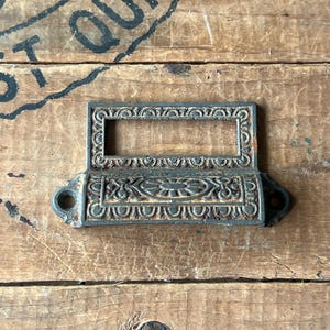 May include: An antique, cast iron drawer pull with a rectangular label holder. The pull has an ornate, embossed design with a central oval and scrollwork. The metal has a weathered, rustic appearance, suggesting age and use. The pull is mounted on a wooden surface.