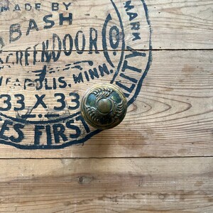 May include: An antique brass doorknob with an ornate floral design, set against a weathered wooden surface. The wood has faded black text from the Bash Door Co. of Minneapolis, Minn. The doorknob is a vintage hardware piece.