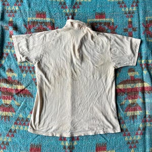 May include: A plain white t-shirt with a collared neckline. The shirt is laying flat on a patterned blue and brown fabric.