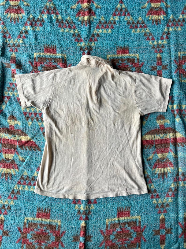 May include: A plain white t-shirt with a collared neckline. The shirt is laying flat on a patterned blue and brown fabric.