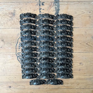 May include: A collection of 30 black cast iron drawer pulls with an ornate design. The pulls are arranged in rows on a wooden surface.