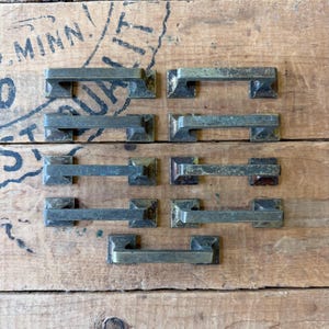 May include: A collection of seven rectangular metal drawer pulls with a weathered, aged brass finish. Each handle features a simple, straight bar design with square mounting plates at both ends, arranged in a grid pattern of three rows.