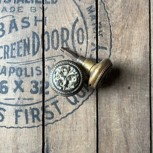May include: Two antique brass door knobs with ornate floral designs. The knobs are on a wooden surface with black lettering that reads "Crystalized Made By Bash Screen Door Co. Indianapolis 6 x 32 S First"