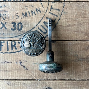 May include: Antique bronze door handle set. The round handle features an embossed design of a vase with plants and stars. The handle has a decorative, ridged base and a long, thin stem with three holes. The set is on a wooden surface.