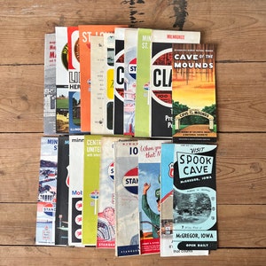 May include: A collection of vintage travel brochures and maps, featuring various destinations. The brochures have colorful covers with text and illustrations, including "Cave of the Mounds" and "Spook Cave". The items are arranged on a wooden surface.
