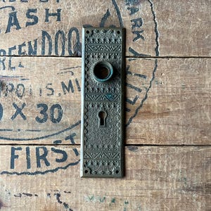 May include: An antique bronze door plate with intricate floral and geometric patterns. The rectangular plate features a keyhole and a circular knob opening. The aged metal has a patina, suggesting its vintage origin. The plate is mounted on a weathered wooden surface.