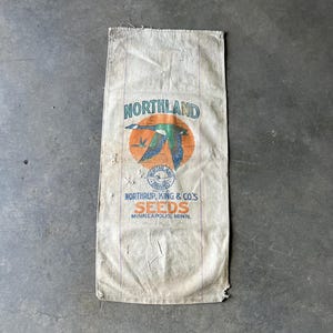Puede incluir: Saco de yute Northland Seeds vintage. El saco presenta un logotipo circular con un pato volador en tonos azules, verdes y naranjas. El texto "NORTHLAND" está sobre el logotipo, con "NORTHRUP, KING & CO'S SEEDS MINNEAPOLIS, MINN." debajo.