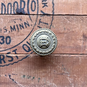May include: A close-up of a brass knob with a decorative design. The knob is set against a wooden background with black ink markings.