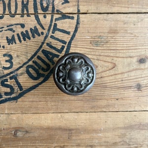 May include: A dark, ornate metal cabinet knob with a floral design. The knob is circular with a raised central element and decorative flourishes around the edge. It rests on a wooden surface.