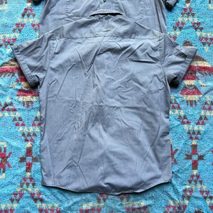 以下が含まれることがあります： ボタンダウンカラーとポケットが付いたグレーの半袖シャツ2枚。シャツは青と茶色の模様の表面に平らに置かれています。