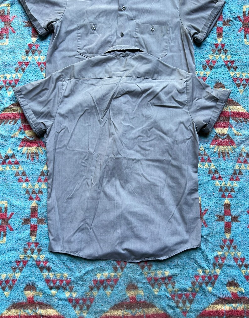 以下が含まれることがあります： ボタンダウンカラーとポケットが付いたグレーの半袖シャツ2枚。シャツは青と茶色の模様の表面に平らに置かれています。