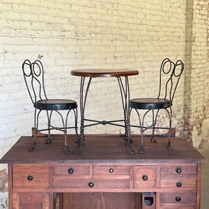 May include: A small bistro set with a round wooden table and two matching chairs. The chairs and table have black metal frames with decorative swirls and a dark brown finish. The chairs have round, black seats.
