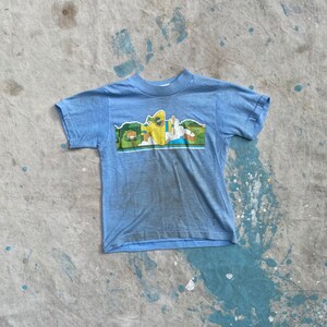 May include: A light blue t-shirt with a cartoon graphic of a yellow plane flying over a green landscape. The plane has a pilot and a passenger in the cockpit.