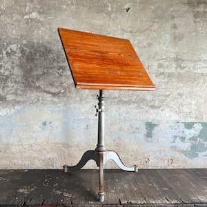 May include: A vintage drafting table with a wooden top and a gray metal base. The table is adjustable and has three legs.