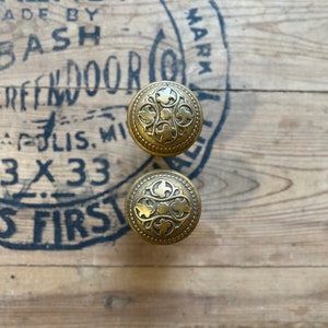 May include: Two gold-toned door knobs with a decorative floral design. The knobs are on a wooden surface.