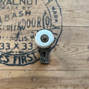 May include: A white porcelain door knob with a brass center, mounted on a black metal plate. The knob is attached to a wooden door with a black painted circle that reads "Bash Walnut Made by Bash Screen Door Co. Indianapolis, IN 33 x 33 Mark"