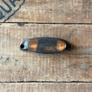 May include: An antique, oval-shaped metal drawer pull with a dark, aged patina. The pull has a central, slightly raised section with a lighter, copper-colored hue, flanked by darker, almost black areas. Small holes are visible on each end for mounting.