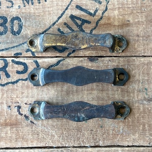 May include: Three antique metal drawer pulls with a weathered, aged appearance. The pulls are a mix of brass and dark metal, with a curved, ergonomic design. They are mounted on a rustic wooden surface, suggesting a vintage or repurposed aesthetic.