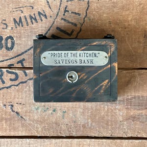May include: A vintage wooden savings bank with a metal plaque that reads "PRIDE OF THE KITCHEN, SAVINGS BANK".