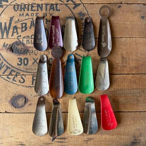May include: A collection of vintage shoe horns in various colors and materials, including metal and plastic. Some shoe horns have text, such as "Sears", "Hush Puppies", and "Caesars Palace". The shoe horns are arranged on a wooden surface.