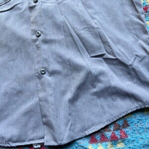 以下が含まれることがあります： 左側にポケットが付いた薄い青色のボタンダウンシャツ。シャツは前面にボタンが2つ見える。