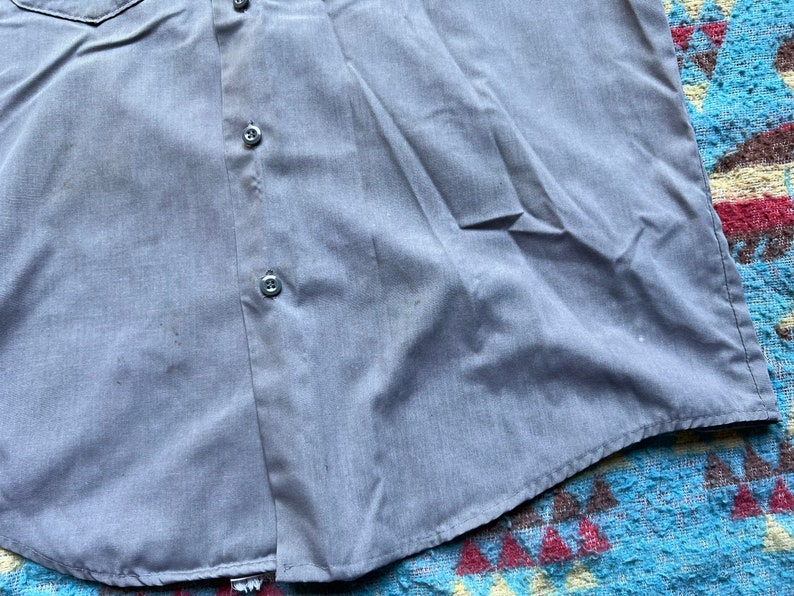 以下が含まれることがあります： 左側にポケットが付いた薄い青色のボタンダウンシャツ。シャツは前面にボタンが2つ見える。