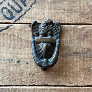 May include: A bronze-colored door knocker featuring an eagle with outstretched wings. The knocker has a curved handle and a decorative design. The word "QUAL" is partially visible in the background, printed in black ink on the wooden surface.