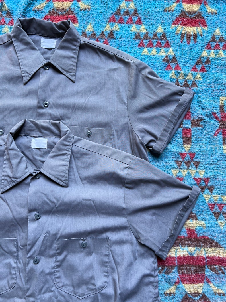 以下が含まれることがあります： 胸にポケットが付いたグレーの半袖ボタンダウンシャツが2枚です。
