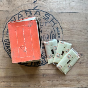 May include: A box of electrical wiring devices with the text "The Sesco Line of Fine Wiring Devices" printed on the side. The box is sitting on a wooden surface with three light beige electrical switch plates.