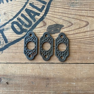 May include: Three antique cast iron door or window hardware pieces with ornate designs. The pieces are dark brown and have a rustic look.