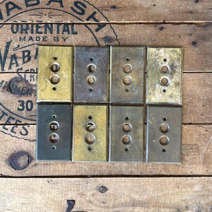 May include: Eight vintage brass and metal light switch covers in various shades of gold, bronze, and silver. Each cover has two circular holes for the switch and two screw holes. The covers are rectangular and arranged in two rows of four.