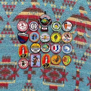 May include: A collection of 20 colorful embroidered patches featuring various scout-related designs. The patches are circular and feature a variety of colors, including red, blue, yellow, green, and white. The patches are arranged in a circular pattern on a blue and brown patterned background.