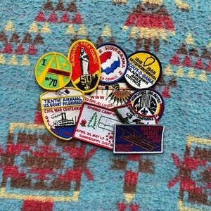 May include: A collection of vintage embroidered patches featuring various designs, including a scout-o-rama event, a civil war centennial, a spring camp, and a Klondike derby. The patches are colorful and feature a variety of text and images.