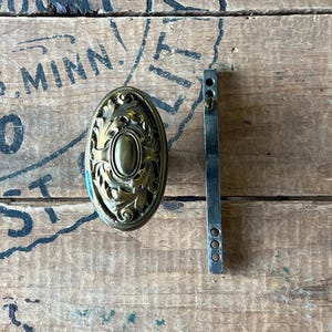May include: An antique brass oval door knob with ornate floral detailing and a matching metal strike plate. The hardware is set against a weathered wooden surface, showcasing a vintage aesthetic. The door knob is approximately 4 inches long.
